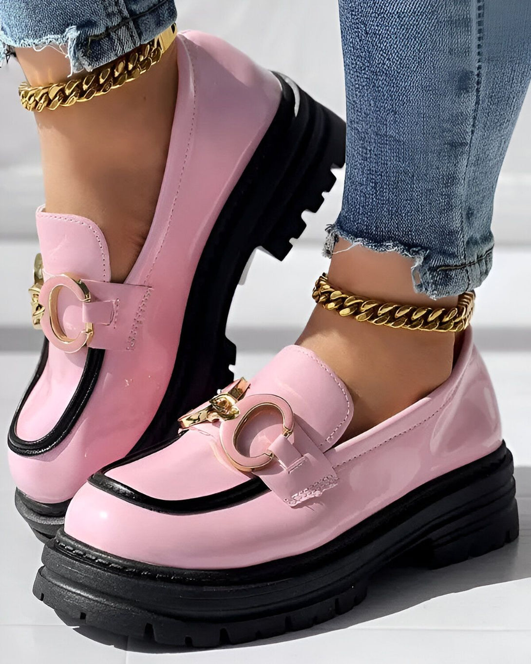 Une femme porte des mocassins roses et noirs avec des semelles épaisses. Elle porte également un jean bleu et des chaussettes blanches. Les mocassins sont en cuir verni et ont une boucle dorée sur le devant.