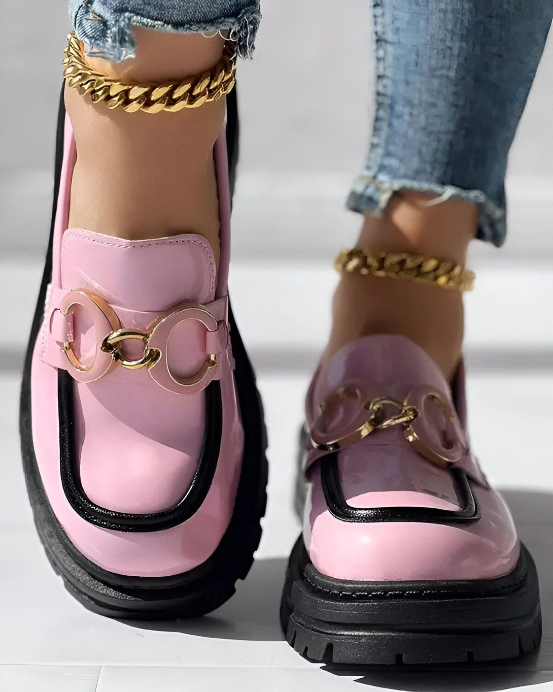 Une femme porte des mocassins roses et noirs avec des chaînes dorées. Elle porte également un jean bleu. Les mocassins sont en cuir verni et ont une semelle épaisse. La femme est debout sur un sol en marbre blanc.
