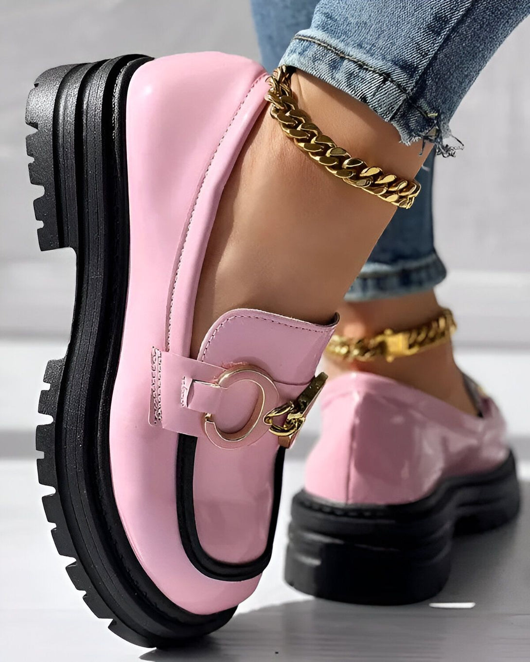Une image d'une paire de chaussures roses et noires avec une boucle dorée. Les chaussures sont portées par une femme qui porte des jeans bleus et un bracelet de cheville doré.
