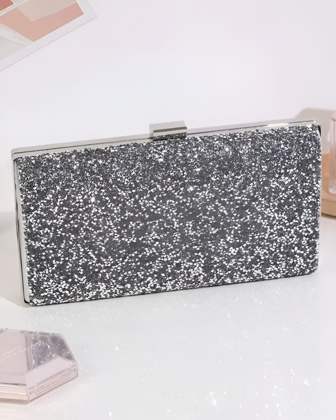 Une image d'une pochette de soirée rectangulaire argentée avec une fermeture à fermoir. La pochette est posée sur une surface blanche.