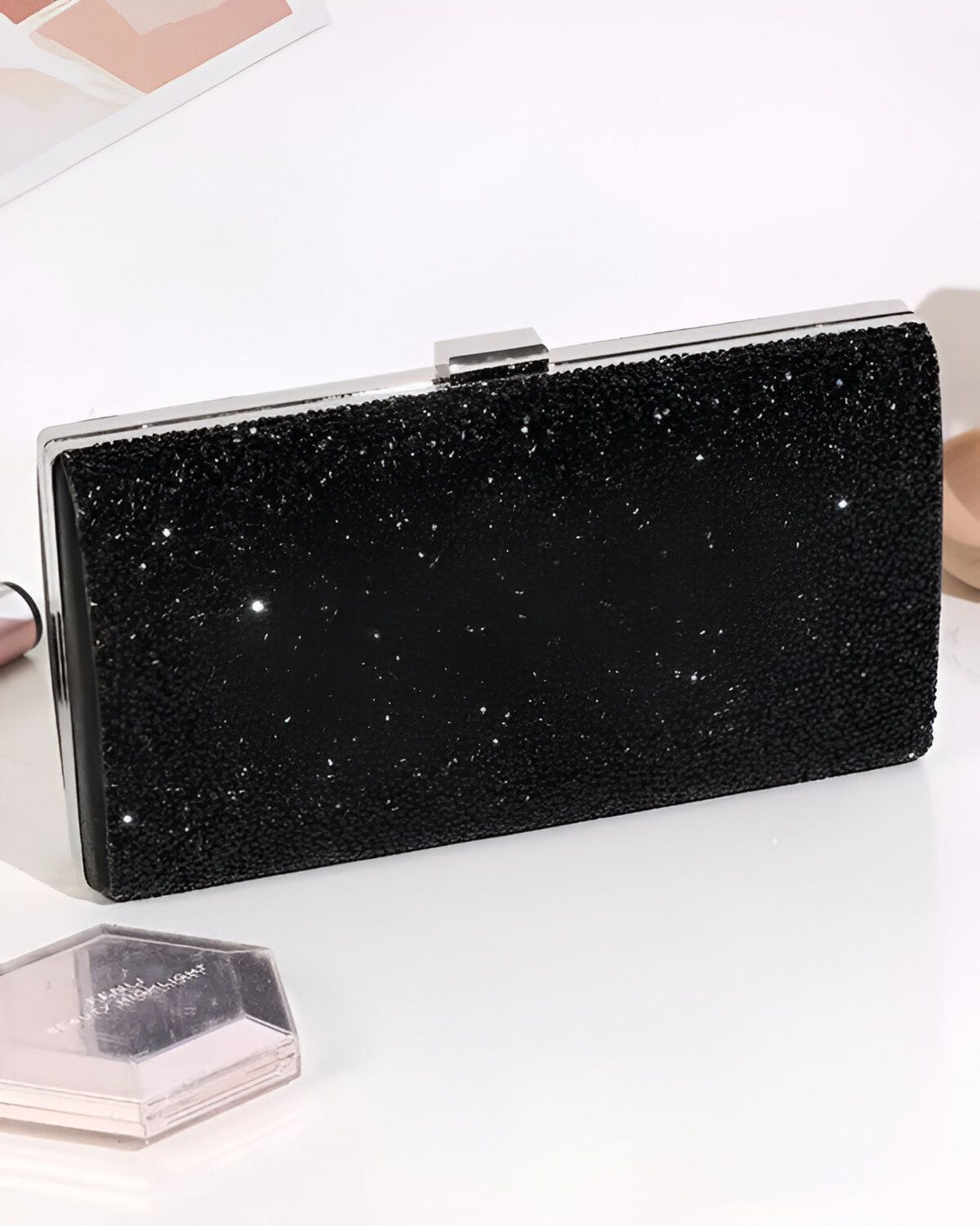 Une image d'une pochette de soirée noire en strass avec une anse argentée. La pochette est posée sur une table blanche à côté d'un poudrier rose et d'un rouge à lèvres.