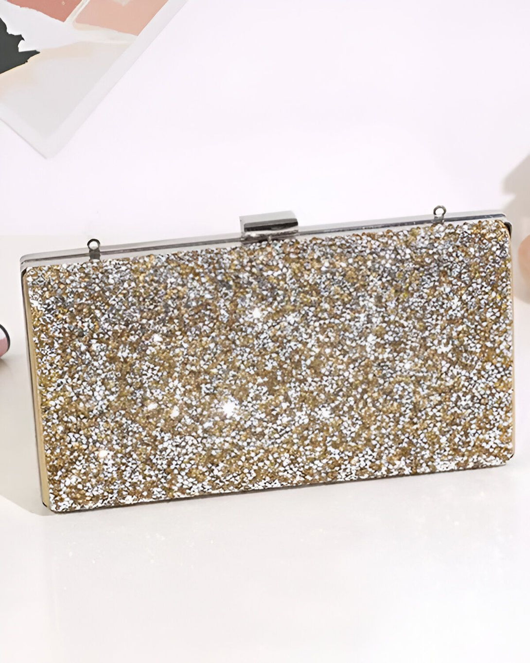 Une image d'une pochette de soirée rigide de couleur or avec une anse argentée. La pochette est recouverte de paillettes et de strass.