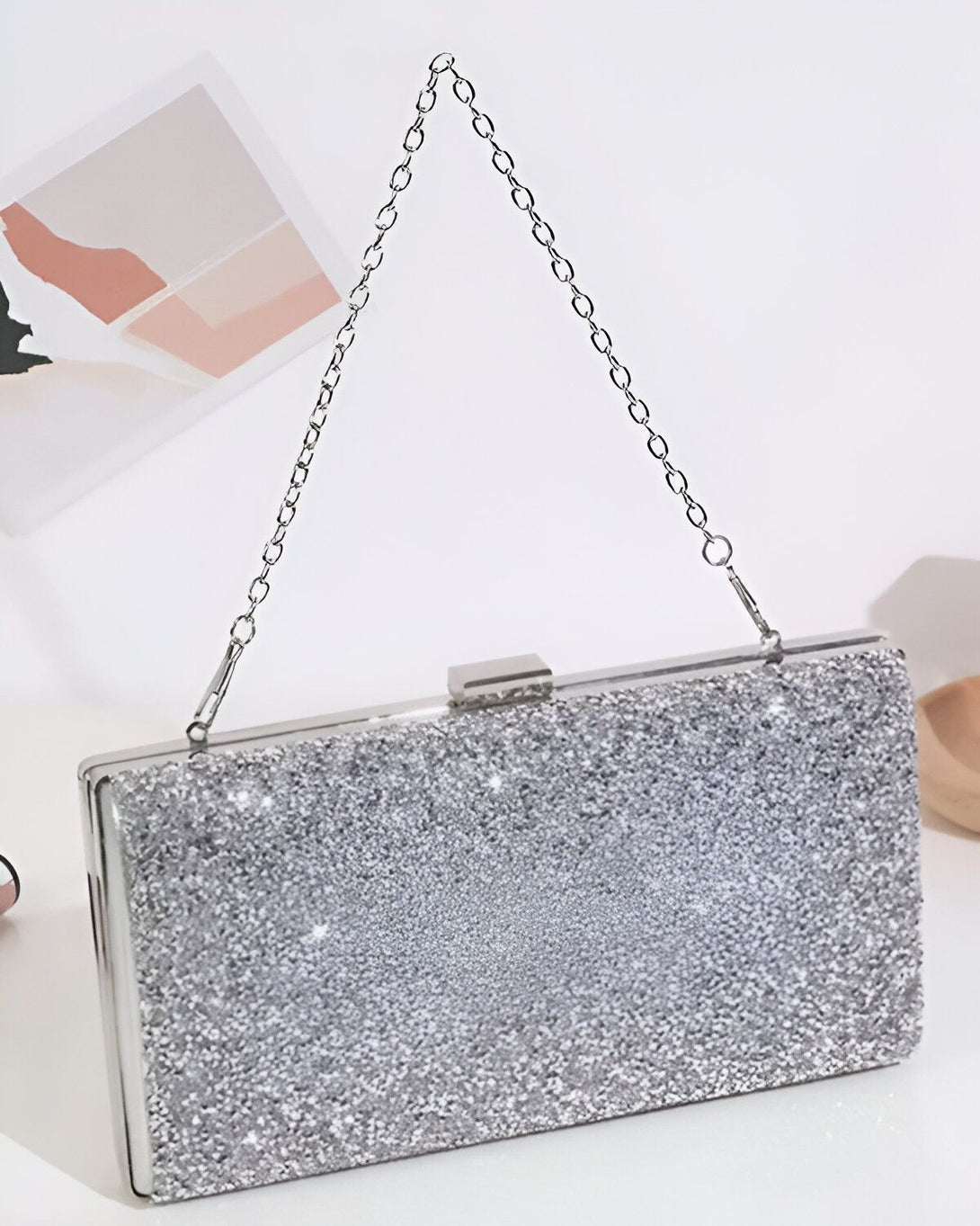 Une image d'une pochette de soirée argentée avec une chaîne. La pochette est ouverte et on peut voir l'intérieur qui est en satin noir. La pochette est posée sur un fond blanc.