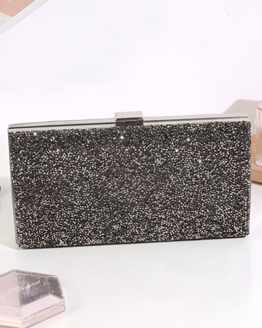 Une image d'une pochette de soirée argentée pailletée. La pochette est rectangulaire et possède une fermeture à fermoir. Elle est posée sur une surface blanche.