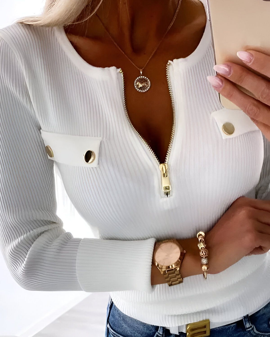 Une femme porte un chemisier blanc moulant avec un décolleté zippé. Le chemisier a des manches longues et des poches sur la poitrine. La femme porte également un jean bleu et une montre en or.