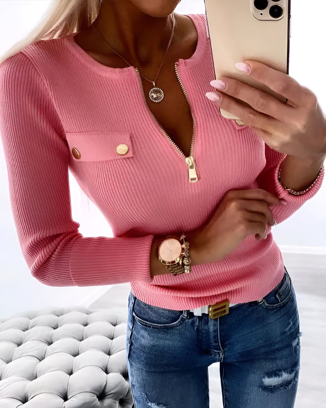 Une femme porte un chemisier rose à manches longues avec un col en V et une fermeture éclair dorée. Elle porte également un jean bleu et un collier avec un pendentif doré.