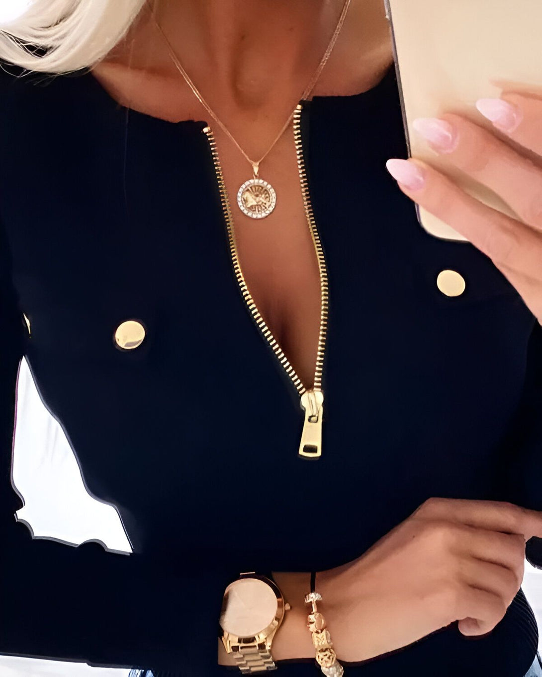 Une femme porte un chemisier noir avec un zip doré sur le devant. Elle porte également un collier avec un pendentif rond en or et des boutons de manchette dorés. Elle tient un téléphone portable dans sa main droite.