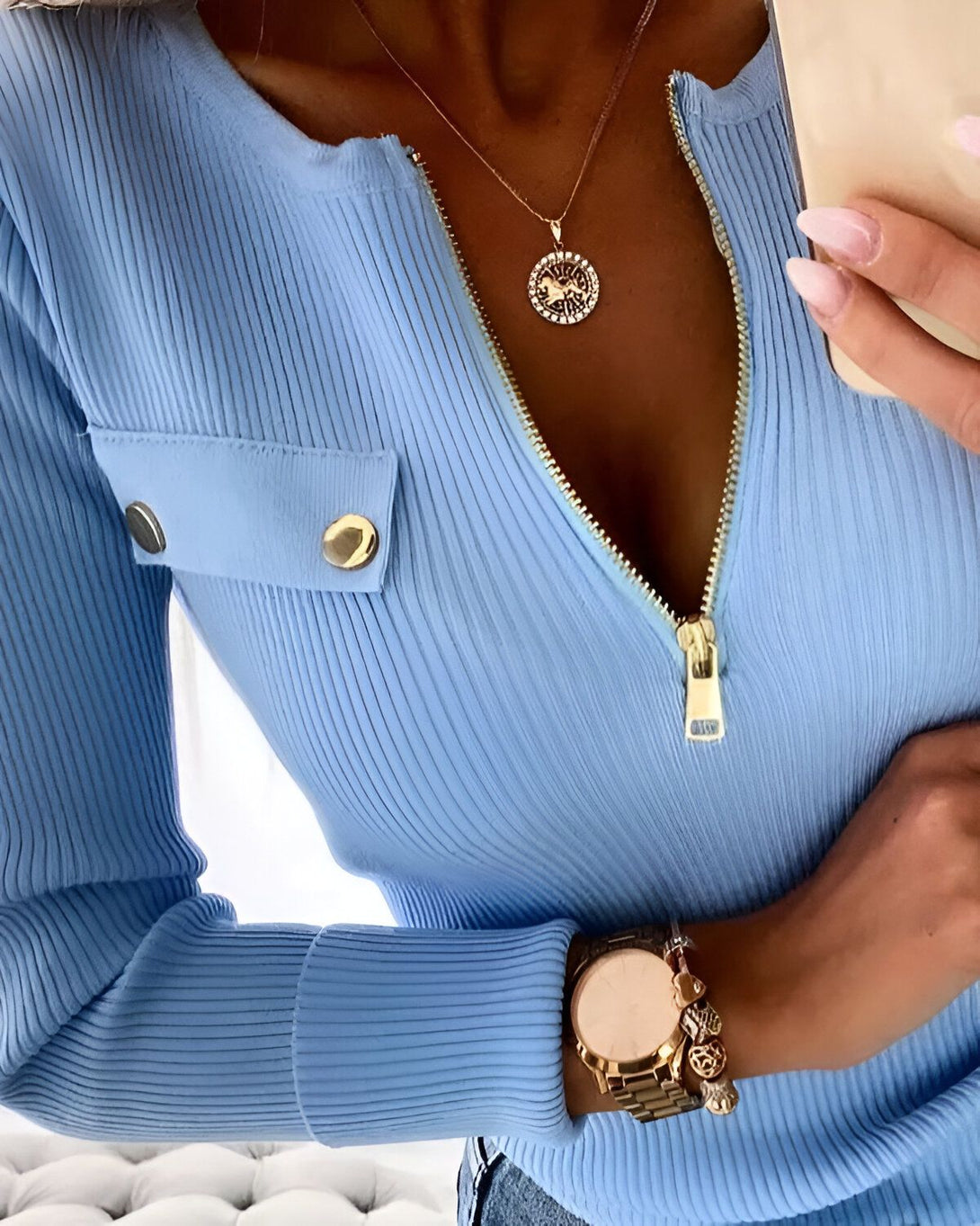 Une femme porte un haut bleu avec une fermeture éclair dorée sur le devant. Elle porte également un collier doré avec un pendentif rond.