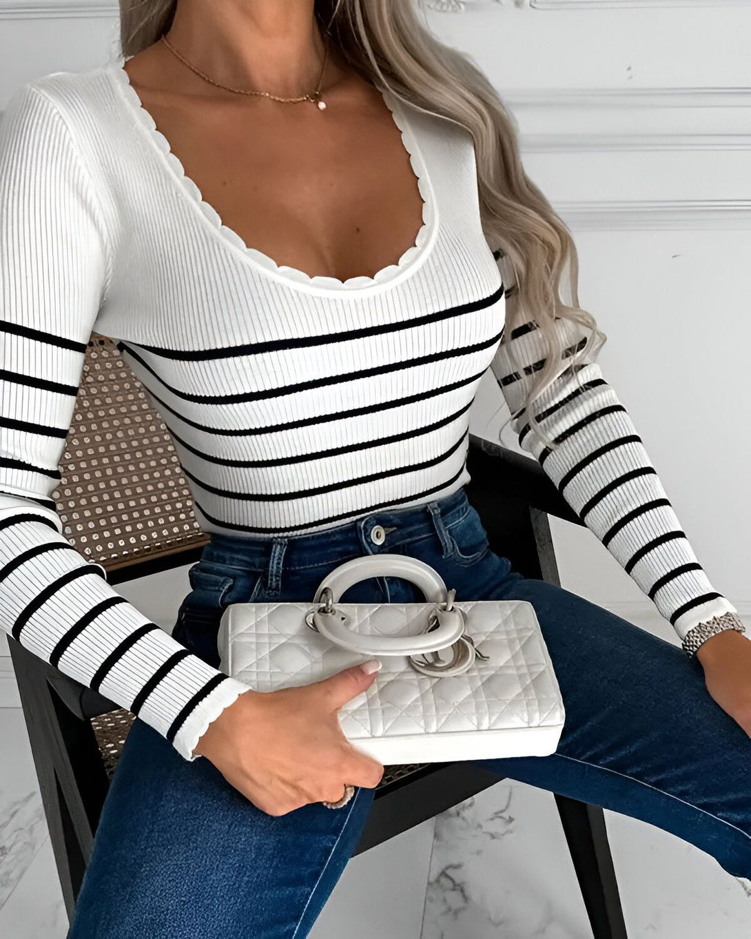 Une femme porte un haut rayé noir et blanc à manches longues et un jean bleu. Elle est assise sur une chaise et tient un sac à main blanc.