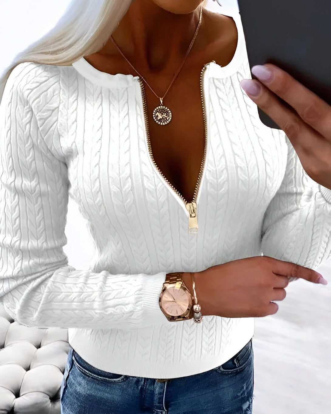 Une femme porte un pull blanc avec une fermeture éclair dorée sur le devant. Le pull est en laine et a un col montant. La femme porte également un jean bleu et une montre en or. Elle a les cheveux blonds et les yeux bleus. Elle est souriante et regarde l'appareil photo.