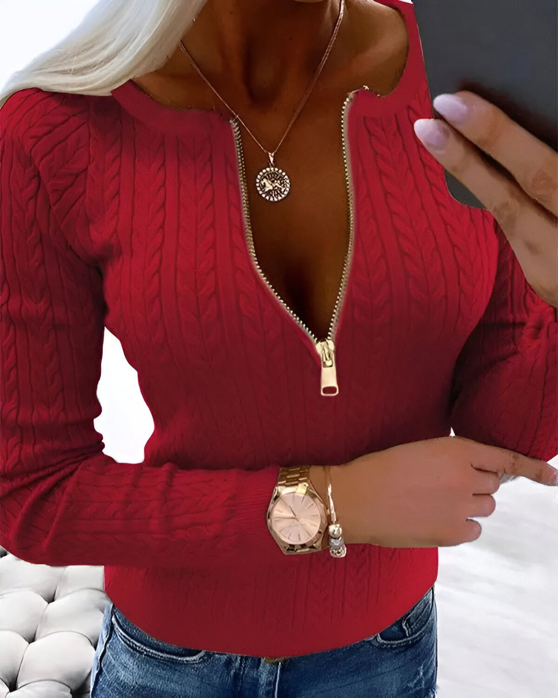 Une femme porte un pull rouge à manches longues avec un zip doré sur le devant. Elle porte également un jean bleu et une montre.