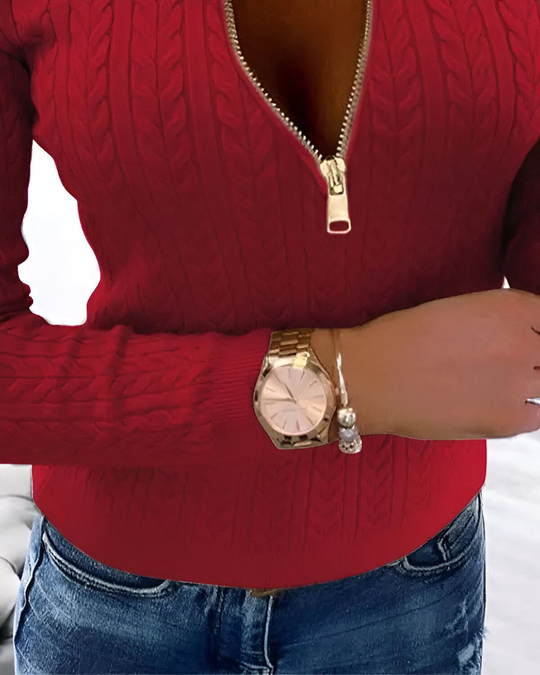 Une femme porte un pull rouge à fermeture éclair et un jean bleu. Elle a une montre en or au poignet gauche.