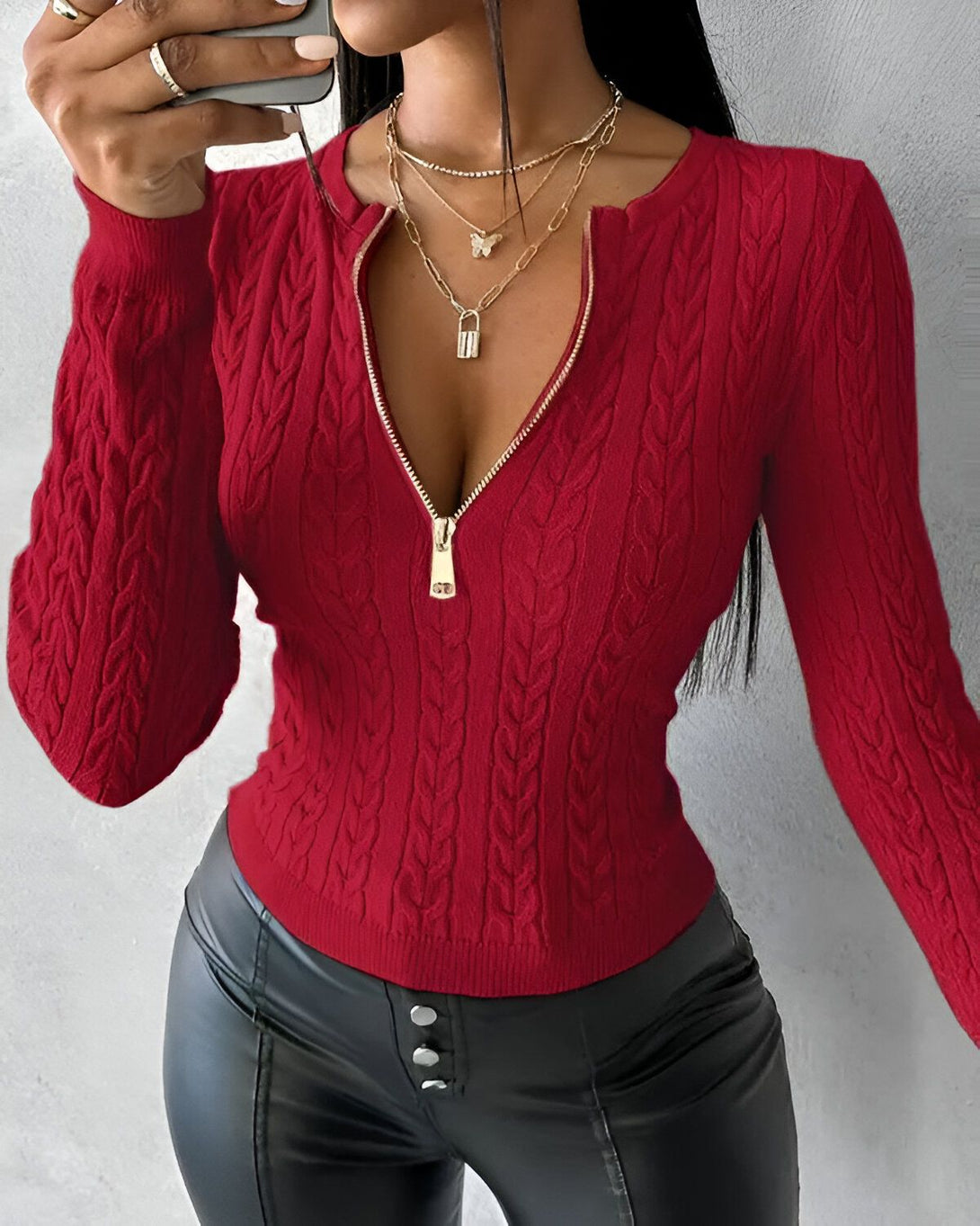 Une femme porte un pull rouge à col montant et à manches longues. Le pull est zippé sur le devant. La femme porte également un collier avec un pendentif en forme de cadenas.