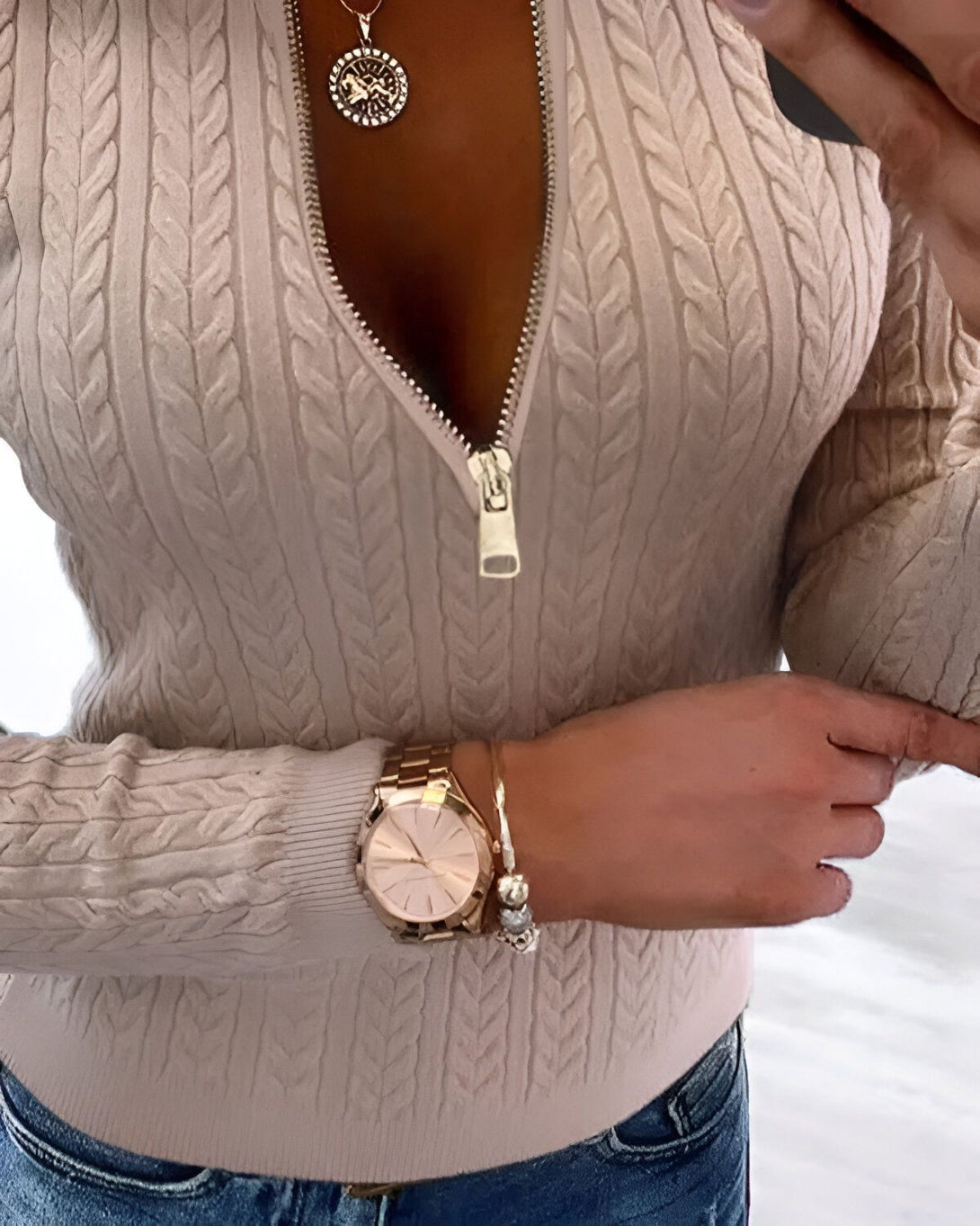 Une femme porte un pull beige avec une fermeture éclair dorée. Elle porte également une montre rose gold et un collier avec un pendentif rond.