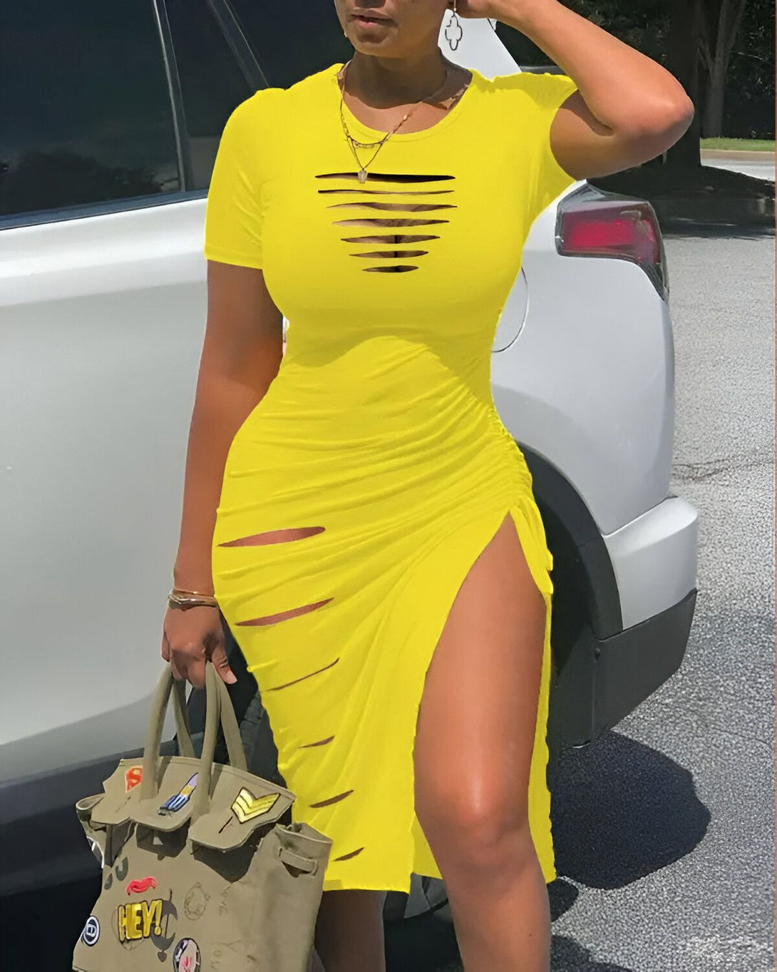 Une femme porte une robe jaune moulante avec des découpes sur le devant et les côtés. La robe est fendue sur le côté droit. La femme est debout à côté d'une voiture et porte un sac à main vert olive.
