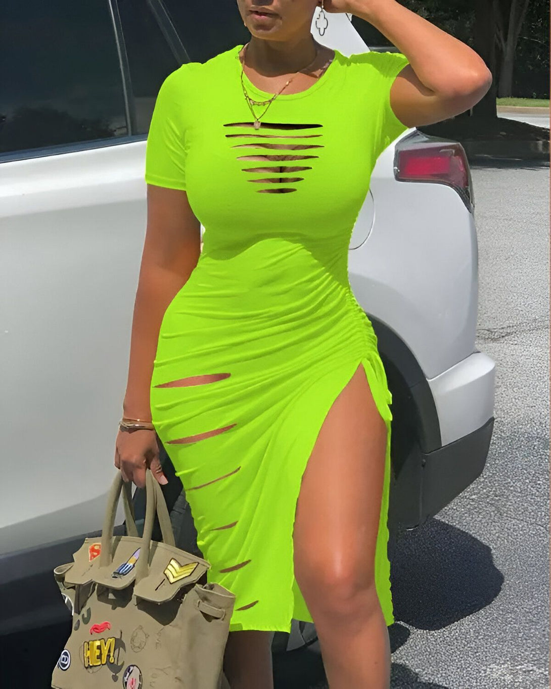 Une femme porte une robe moulante verte fluo avec des découpes sur le devant et une fente sur le côté. Elle porte également un sac à main vert kaki et des talons hauts noirs.