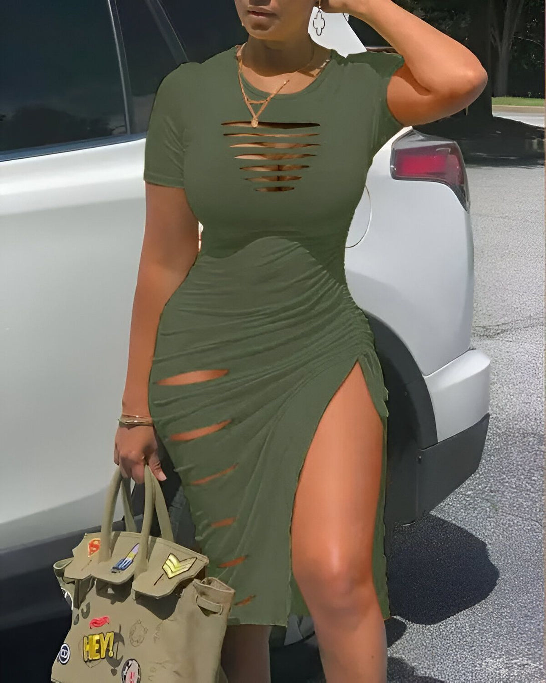 Une femme porte une robe verte moulante avec des découpes sur le devant et une fente sur le côté. Elle est également porte une paire d'escarpins noirs et un sac à main vert.