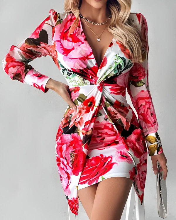 Une femme porte une robe moulante à fleurs avec un décolleté en V et des manches longues. La robe est rouge, blanche et verte. La femme est blonde et porte un collier. Elle est debout dans une pièce blanche et regarde l'appareil photo.