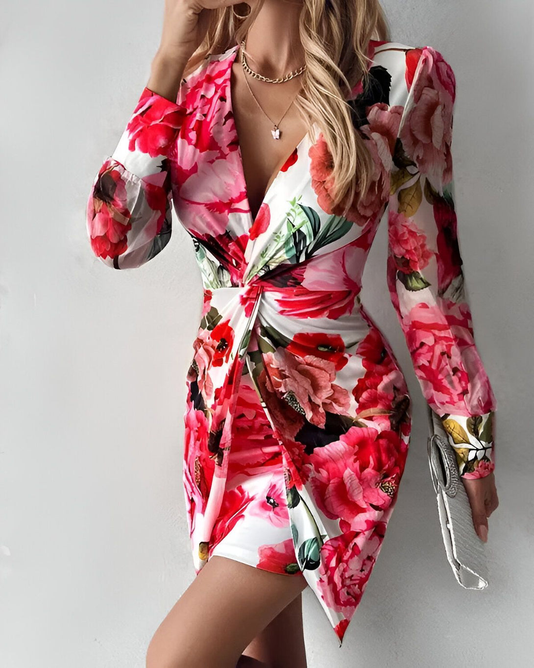 Une femme porte une robe courte à fleurs avec un décolleté en V et des manches longues. La robe est blanche avec des fleurs roses et rouges. La femme est blonde et porte un collier avec un pendentif papillon. Elle porte également un sac à main blanc.