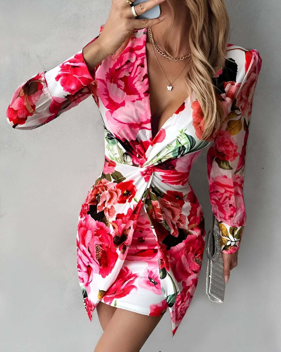 Une femme porte une robe moulante à fleurs avec un décolleté en V et des manches longues. La robe est rose et blanche avec des fleurs rouges et vertes. La femme est blonde et porte un collier avec un pendentif en forme de papillon. Elle porte également des talons hauts argentés et une pochette argentée.