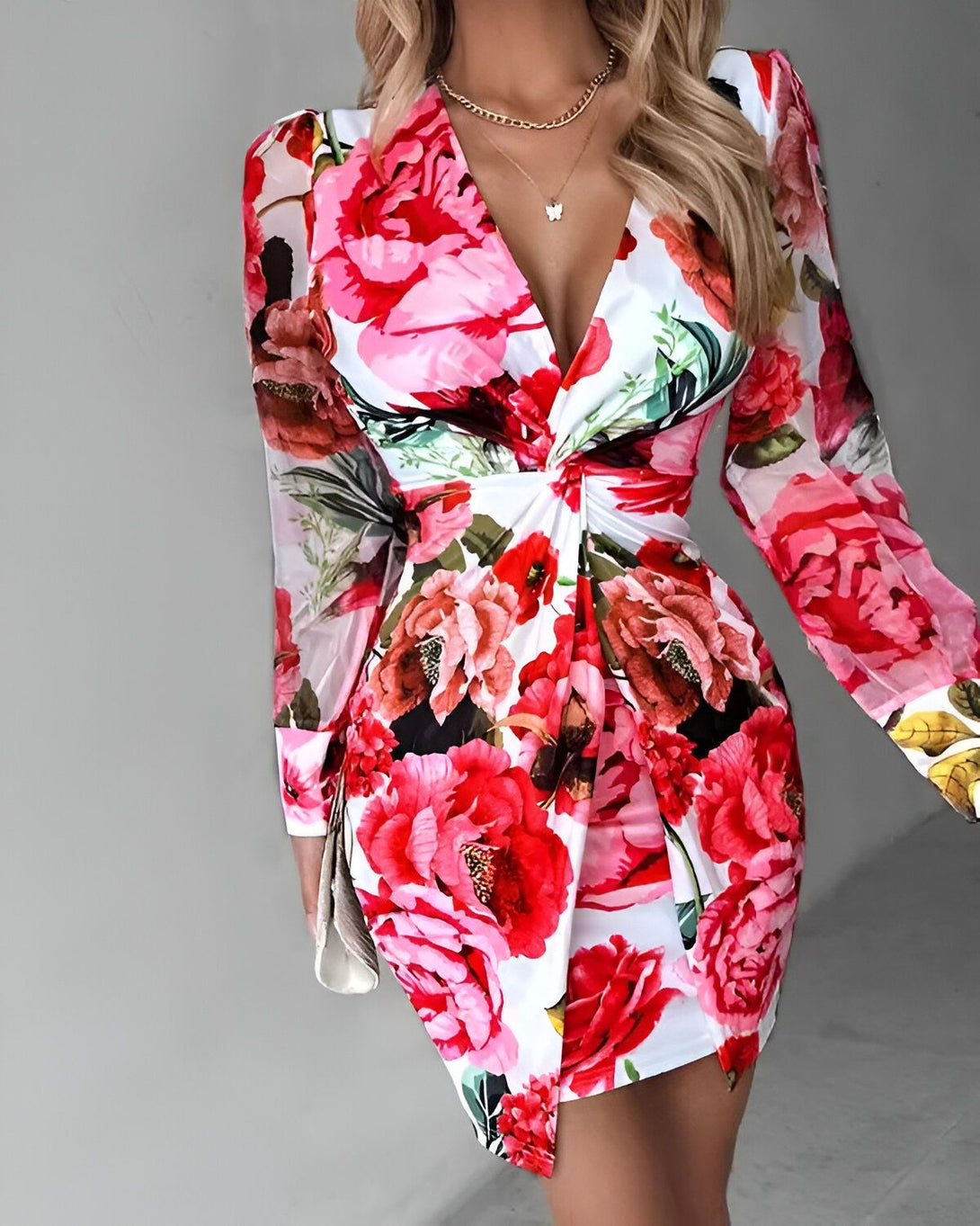 Une femme porte une robe blanche à fleurs rouges et roses. La robe est courte et cintrée, avec un décolleté en V et des manches longues. La femme est blonde et porte un collier avec un pendentif en forme de papillon.