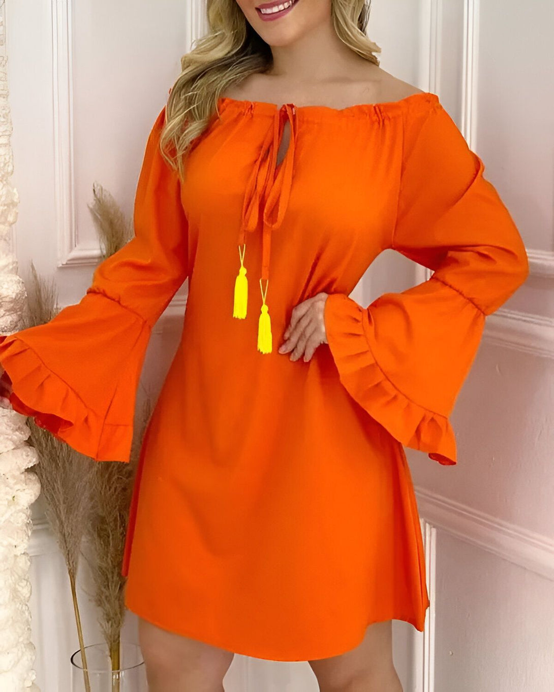 Une jeune femme porte une robe orange à épaules dénudées. La robe a des manches longues et évasées. La femme a les cheveux longs et blonds et elle sourit.
