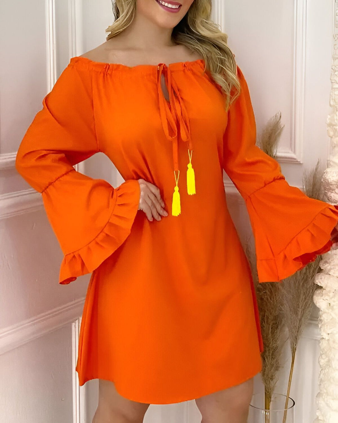 Une jeune femme porte une robe orange à épaules dénudées. La robe a des manches longues et évasées. La femme est souriante et a les cheveux longs et blonds. Elle porte des boucles d'oreilles jaunes et un bracelet jaune.