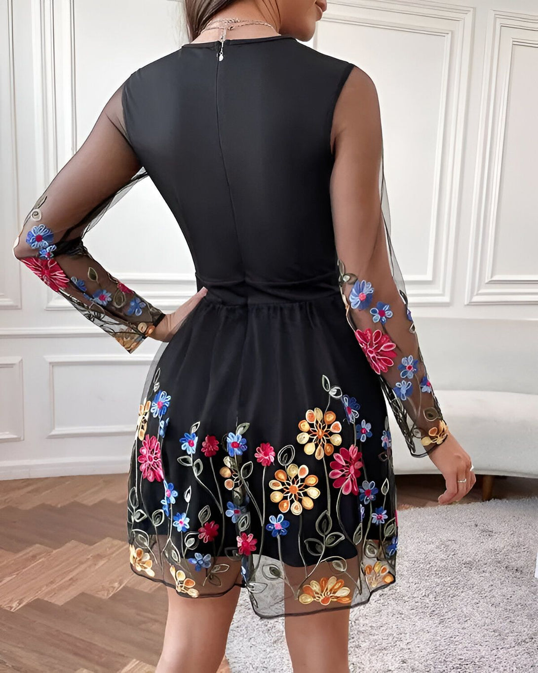 Une femme porte une robe noire en dentelle brodée de fleurs colorées. La robe est courte et évasée, avec des manches longues et un col rond. La femme est vue de dos.