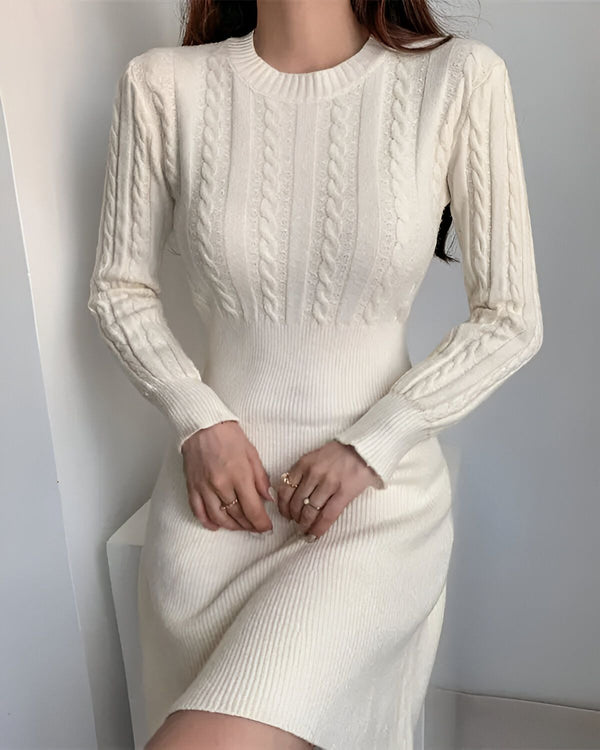 Une jeune femme porte une robe pull blanche en laine torsadée. La robe est mi-longue et possède des manches longues. La femme est vue de face et elle a les cheveux longs et noirs. Elle porte également des bagues aux doigts.