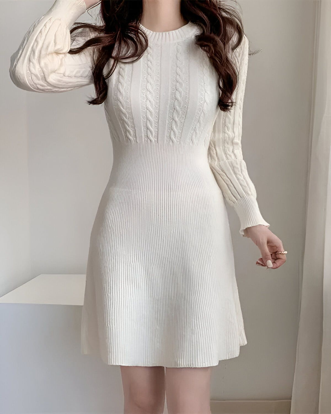 Une jeune femme porte une robe blanche en laine. La robe est à manches longues et a un col rond. La femme a les cheveux longs et noirs et elle est maquillée de façon naturelle. Elle se tient debout dans une pièce blanche et regarde l'appareil photo.
