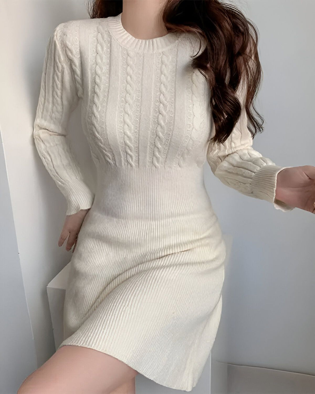 Une jeune femme porte une robe blanche en laine torsadée. La robe est ajustée et a une jupe évasée. La femme a les cheveux longs et bruns et elle est maquillée de façon naturelle. Elle est debout dans une pièce blanche et elle regarde l'appareil photo.
