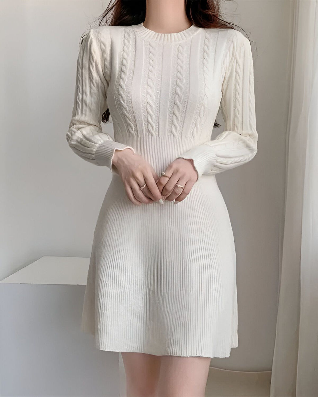 Une jeune femme porte une robe blanche en laine. La robe est à manches longues et a un col rond. La femme a les cheveux longs et noirs et elle est maquillée de façon naturelle. Elle porte des bagues aux doigts.