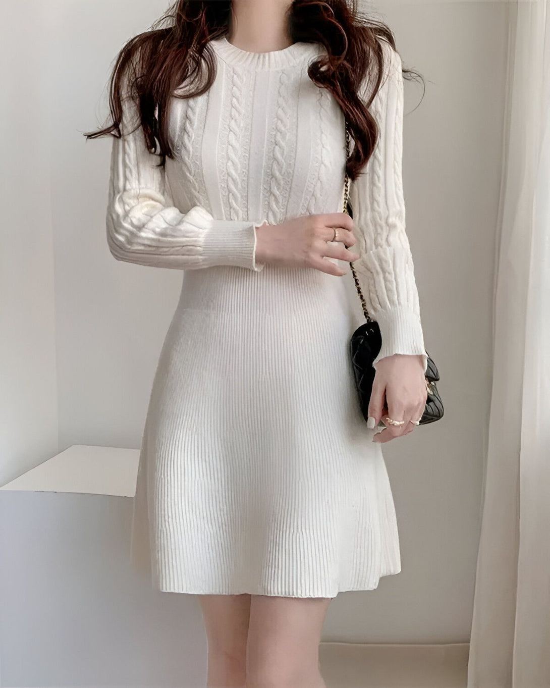 Une jeune femme porte une robe blanche en laine torsadée. La robe est à manches longues et a une jupe évasée. La femme a les cheveux longs et bruns et porte un sac à main noir.