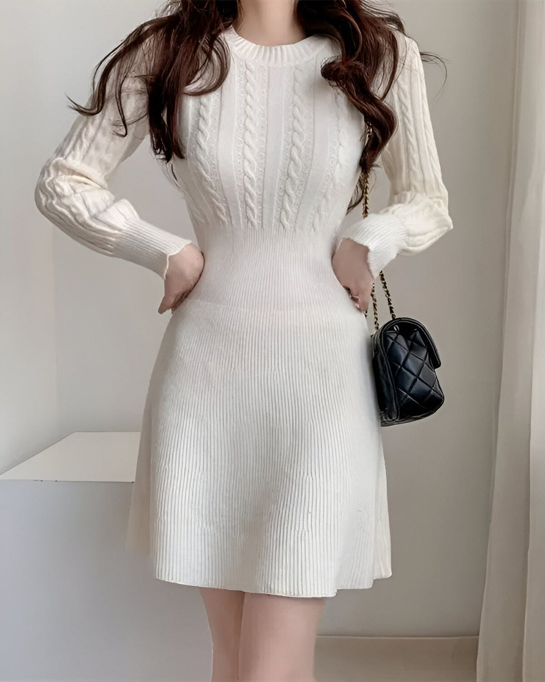 Une jeune femme porte une robe blanche en laine torsadée. La robe est ajustée à la taille et évasée au niveau des hanches. Elle a des manches longues et un col rond. La femme porte également un sac à main noir.