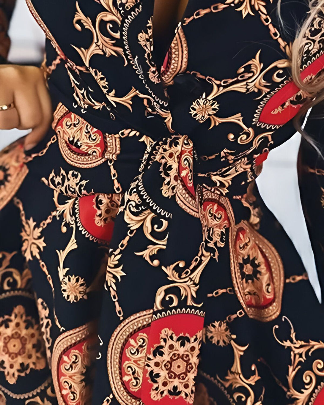 Une femme porte une robe noire avec des motifs baroques rouges et dorés. La robe est cintrée à la taille par une ceinture à nouer.