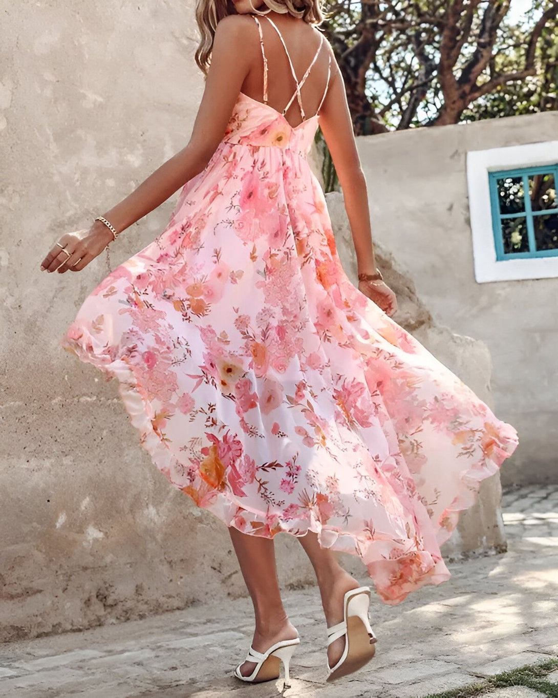 Une femme porte une robe longue rose à fines bretelles. La robe est imprimée de fleurs colorées. La femme est vue de dos. Elle a les cheveux longs et blonds. Elle porte des sandales à talons hauts blanches.