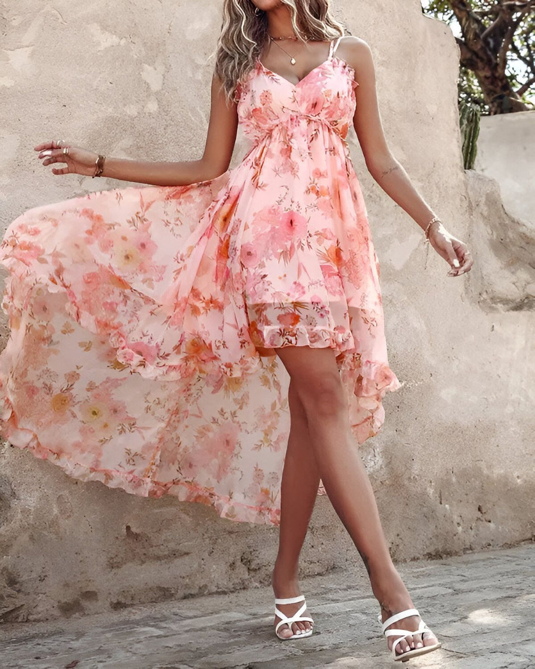 Une femme porte une robe longue rose à motifs floraux. La robe est fendue sur le devant et nouée au niveau de la taille. Elle porte des sandales blanches à talons.