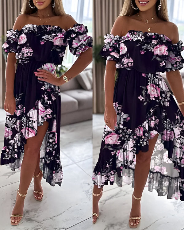 Une femme porte une robe noire à fleurs. La robe est courte devant et longue derrière. Elle a des volants sur les épaules et un décolleté en cœur. La femme est debout et porte des talons hauts.