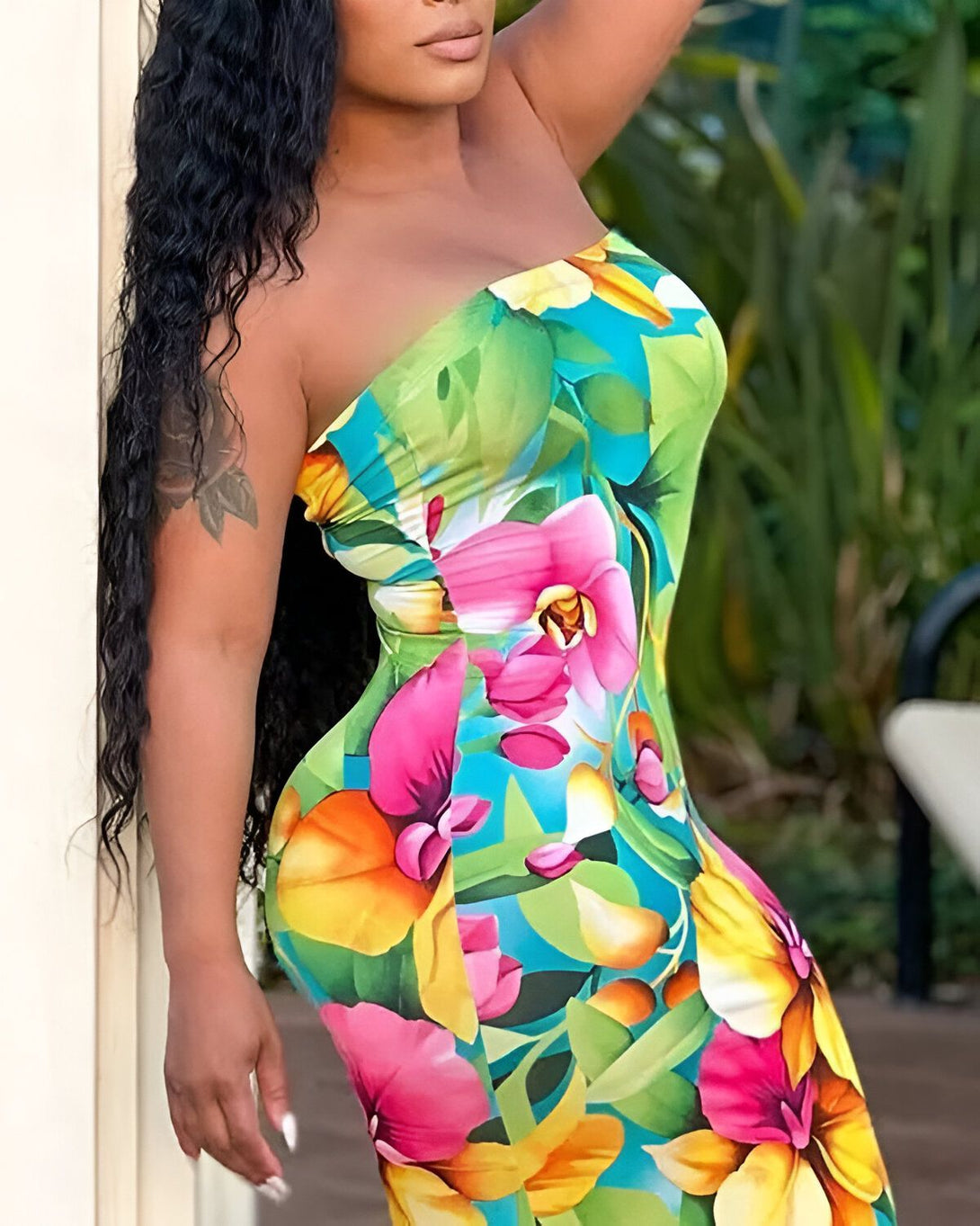 Une femme porte une robe moulante sans bretelles à motifs de fleurs tropicales. La robe est multicolore, avec des fleurs roses, jaunes, vertes et bleues. La femme est debout, adossée à un pilier, et regarde vers l'appareil photo. Elle a les cheveux longs et noirs et porte du maquillage.