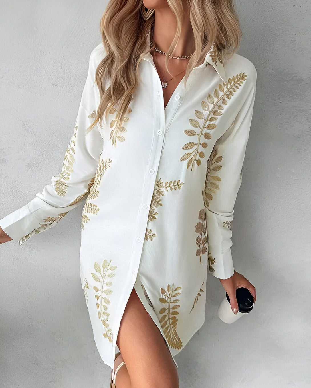 Une jeune femme porte une chemise blanche à manches longues et à col montant. La chemise est ouverte sur le devant et l'on peut voir que la femme porte un soutien-gorge noir. La chemise est également fendue sur les côtés, ce qui laisse entrevoir les jambes de la femme. La femme porte des talons hauts noirs et un collier doré. Elle a les cheveux longs et blonds et elle est maquillée de façon naturelle.