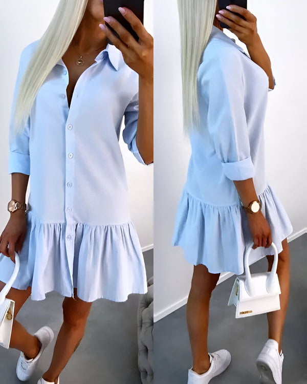 Une jeune femme porte une robe chemise bleue courte à volants. Elle est cintrée à la taille et possède un col montant. La robe est ouverte sur le devant et fermée par des boutons. La jeune femme porte des baskets blanches et un sac à main blanc.