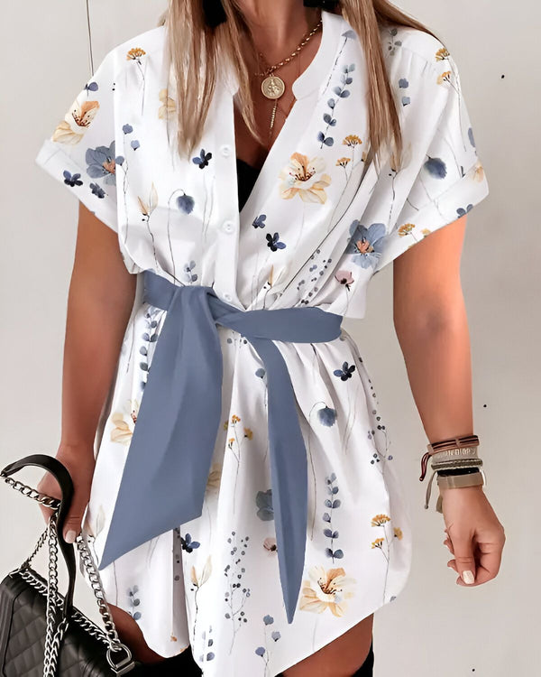 Une femme porte une robe blanche à fleurs avec une ceinture bleue. La robe est courte et a des manches courtes. La femme a les cheveux longs et blonds et porte un collier. Elle porte également un sac à main noir.