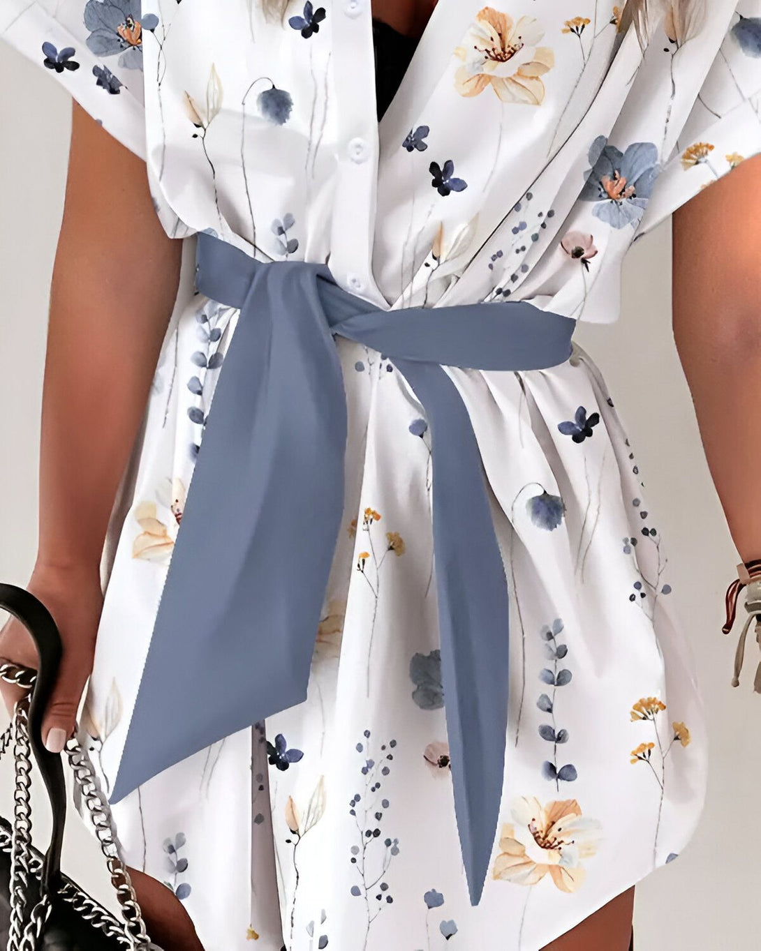 Une jeune femme porte une robe chemise blanche à fleurs bleues et jaunes. La robe est ceinturée d'un ruban bleu.
