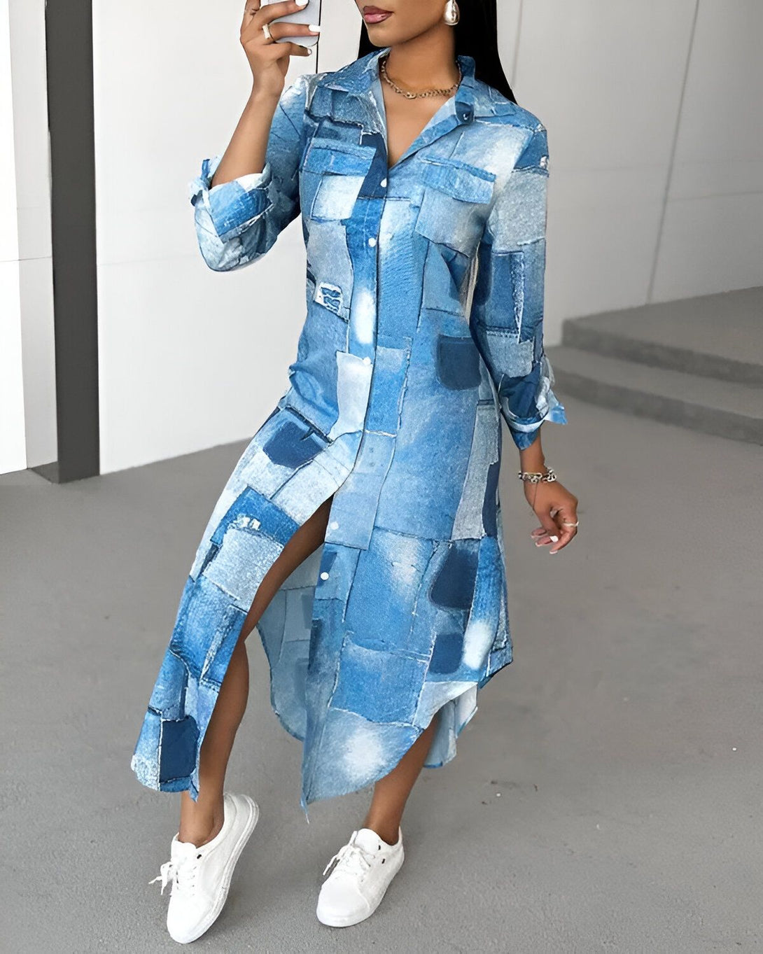 Une femme porte une robe chemise longue en jean bleu. La robe est ouverte sur le devant et fendue sur les côtés. Elle porte des baskets blanches et un collier.