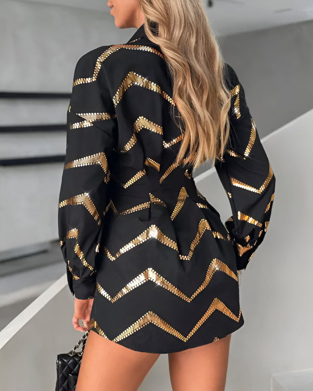 Une femme porte une robe noire et or. La robe est courte et cintrée, avec un col montant et des manches longues. Elle est également dotée d'un motif à chevrons doré. La femme est blonde et porte un sac à main noir. Elle se tient debout dans un escalier, dos à la caméra.