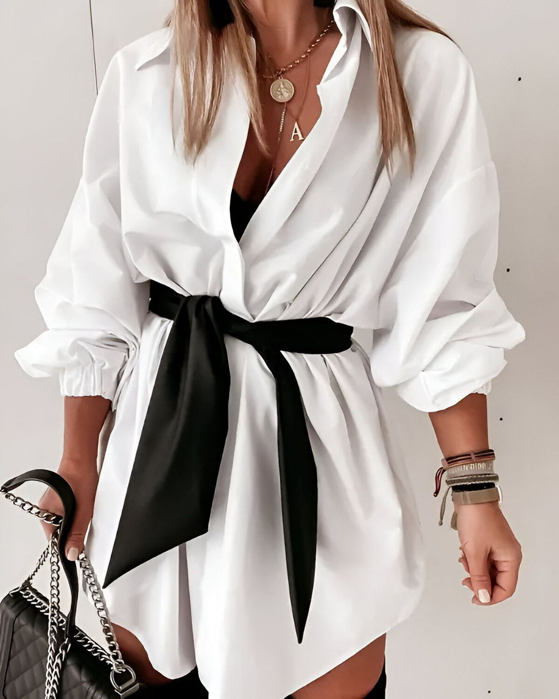 Une femme porte une chemise blanche oversize avec une ceinture noire nouée à la taille. Elle porte également un collier avec un pendentif en forme de A, des bracelets et une montre. Elle tient un sac à main noir à la main.