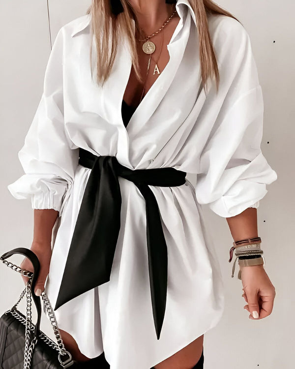 Une femme porte une chemise blanche oversize avec une ceinture noire nouée à la taille. Elle porte également un collier avec un pendentif en forme de A, des bracelets et une montre. Elle tient un sac à main noir à la main.