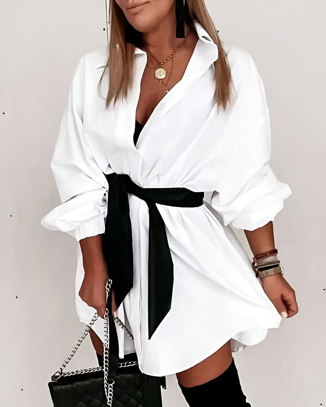 Une femme porte une chemise blanche oversize avec une ceinture noire nouée à la taille. Elle porte également des bottes noires et un collier avec un pendentif rond. Elle a les cheveux longs et blonds et un maquillage léger. Elle est debout devant un mur blanc.