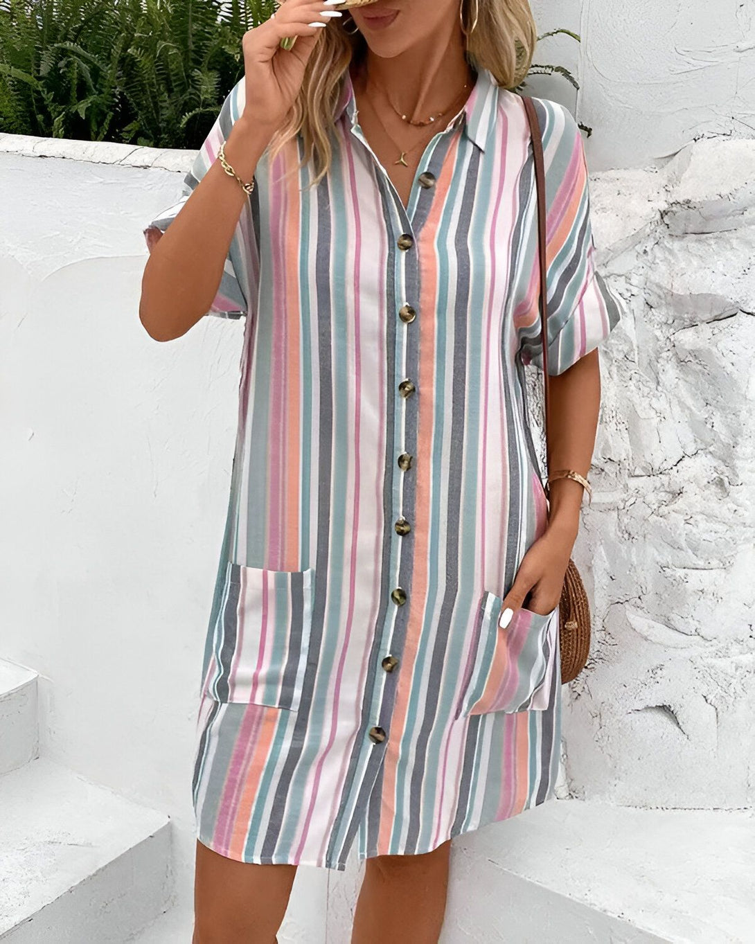 Une femme porte une robe chemise à rayures verticales multicolores. La robe est courte, avec des manches courtes et un col montant. Elle est ouverte sur le devant et fermée par des boutons. La femme est blonde et porte un collier et un sac à main en osier. Elle se tient debout devant un mur blanc.