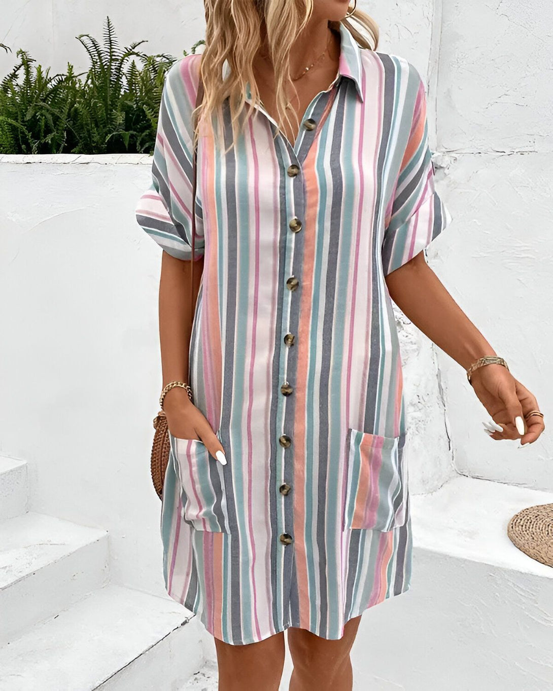 Une femme porte une robe chemise à rayures colorée. La robe est mi-longue et possède des manches courtes. Elle est ouverte sur le devant et possède des boutons. La femme est blonde et porte un collier et des bracelets. Elle a les ongles manucurés.